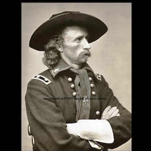 Peut inclure: Un portrait en noir et blanc d'un homme en uniforme militaire. Il porte un chapeau noir à large bord et une écharpe sombre autour du cou. Il a une moustache et les cheveux longs. Il est debout, les bras croisés.