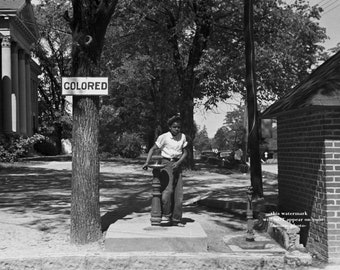 May include: A black and white photo of a young Black boy standing at a water fountain with a sign that reads "COLORED" on a tree behind him.
