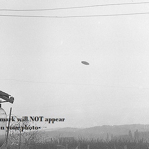 May include: Black and white photograph of a flying saucer hovering in the sky. The image includes a building, power lines, and a distant landscape. The text "-watermark will NOT appear on your photo-" is visible.