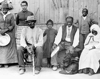 May include: A black and white photograph of a group of people, including a woman holding a plate, a man with a cane, a woman with a shawl, and a young girl. The group is standing in front of a wooden building.