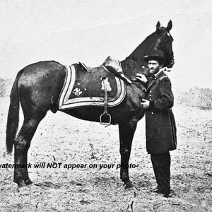 Op de afbeelding: Een zwart-witfoto van een man in militair uniform die naast een zwart paard staat. Het paard draagt een zadel en een hoofdstel. De man houdt de teugels van het paard vast.