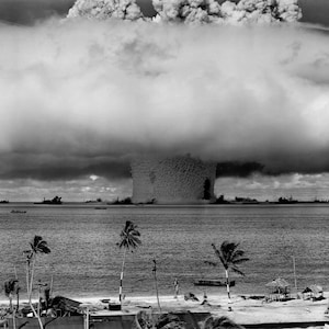 Puede incluir: Una fotografía en blanco y negro de una nube en forma de hongo que se eleva desde el océano. La nube es grande y ondulante, y está rodeada de nubes oscuras y tormentosas. El océano está tranquilo y quieto, y hay palmeras en primer plano.