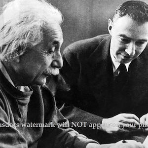 May include: Black and white photo of Albert Einstein and another man sitting at a table. Einstein is on the left, wearing a sweater and looking to the right. The other man is on the right, wearing a suit and tie, and looking down at a piece of paper.