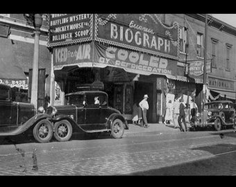 May include: A black and white photo of a movie theater called "Biograph" with a sign that says "Cooled by Refrigeration". There are two cars parked in front of the theater and people walking on the sidewalk.