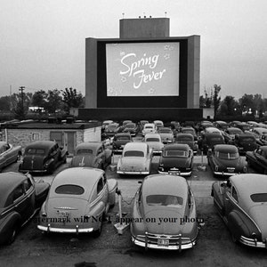 May include: A black and white photograph of a drive-in cinema with a large screen displaying the film "Spring Fever". The screen is surrounded by a white frame. The cinema is full of cars, all facing the screen. The cars are parked in rows, and the people in the cars are watching the film.