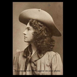 May include: A sepia-toned portrait of a woman with curly hair wearing a wide-brimmed hat with a star on the front. She is wearing a collared shirt with a bow tie.