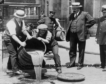 May include: A group of men in early 20th century clothing stand on a city street. One man pours liquid from a barrel into a sewer drain. The men are likely involved in a prohibition-era liquor raid.
