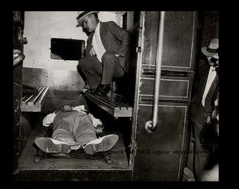 May include: A black and white photograph of a crime scene. A man in a suit and hat is standing over a body lying on the floor. The body is covered in a white sheet. The scene is dark and shadowy.