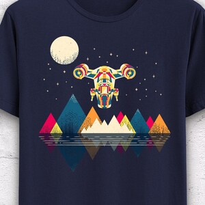 Puede incluir: Camiseta azul marino con un gr&aacute;fico colorido de una nave espacial volando sobre monta&ntilde;as geom&eacute;tricas y un lago reflectante, bajo una luna llena y un cielo estrellado. El dise&ntilde;o incorpora tonos rojos, amarillos, azules y verdes.