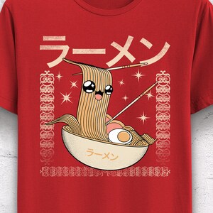 May include: Red t-shirt featuring a cartoon ramen bowl with a smiling face, chopsticks, and the Japanese word for ramen. The design includes a border with decorative elements and stars. The shirt is hanging on a white hanger.