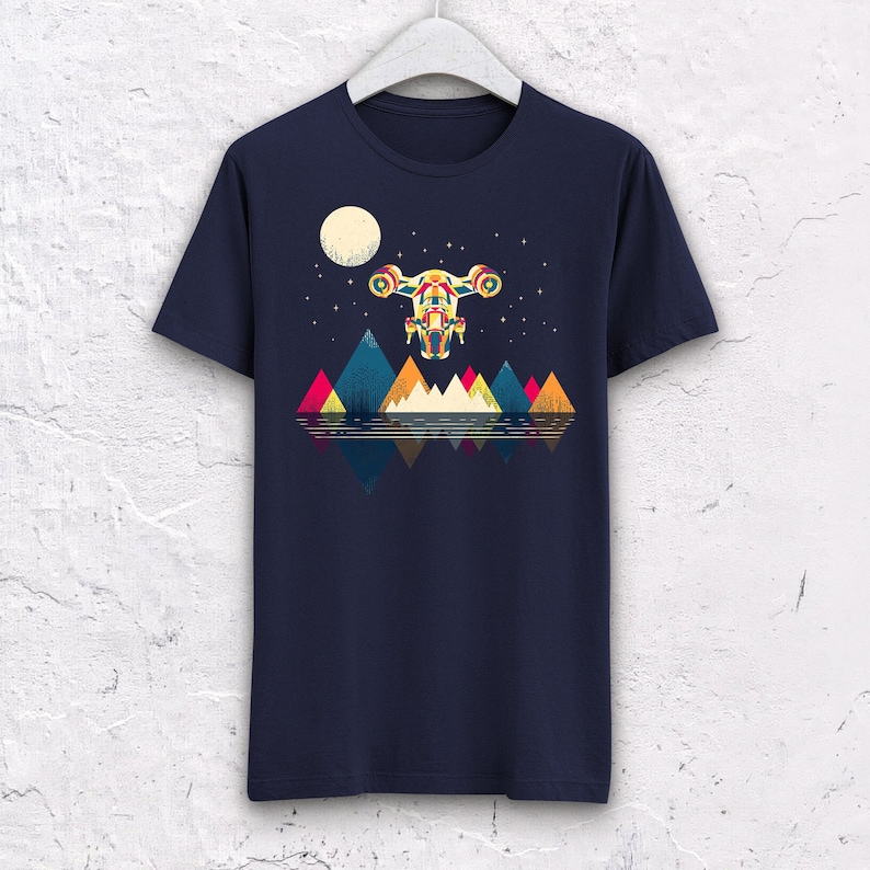 Puede incluir: Camiseta azul marino con un gr&aacute;fico colorido de una nave espacial volando sobre monta&ntilde;as geom&eacute;tricas y un lago reflectante, bajo una luna llena y un cielo estrellado. El dise&ntilde;o incorpora tonos rojos, amarillos, azules y verdes.