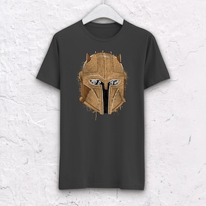 May include: A dark gray t-shirt with a brown and gold illustration of a Mandalorian helmet. The helmet has a visor with the text "This is the way" written on it.