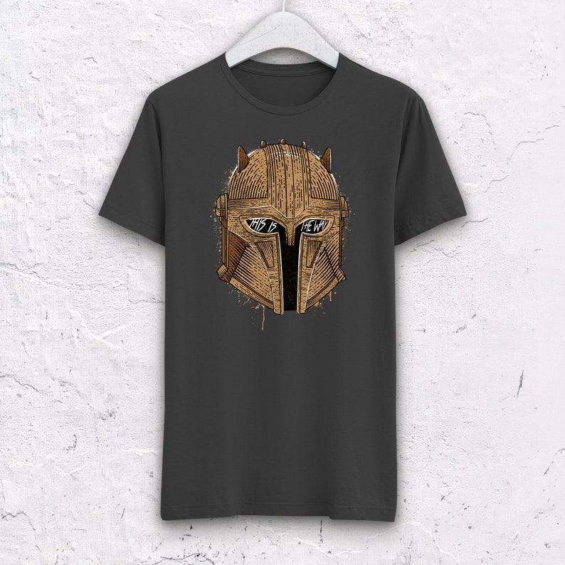 May include: A dark gray t-shirt with a brown and gold illustration of a Mandalorian helmet. The helmet has a visor with the text "This is the way" written on it.