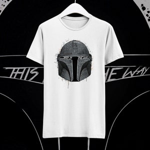 May include: White t-shirt with a black and white illustration of a Mandalorian helmet. The helmet has a splattered paint effect and the text "This is the way" is written on the helmet.