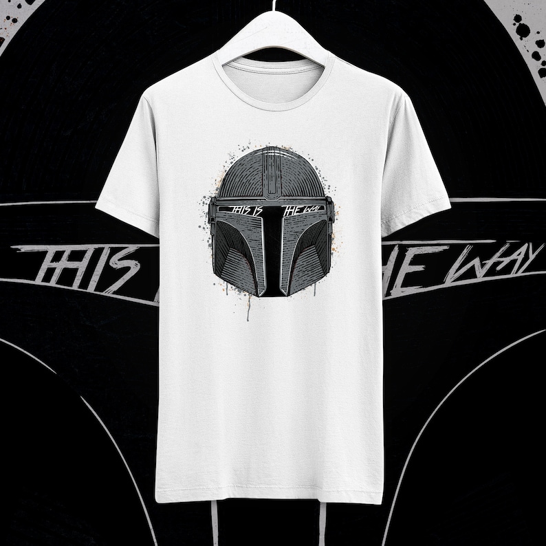 May include: White t-shirt with a black and white illustration of a Mandalorian helmet. The helmet has a splattered paint effect and the text "This is the way" is written on the helmet.