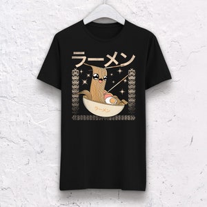 May include: Black t-shirt featuring a cartoon ramen bowl design. The illustration includes a smiling noodle character, chopsticks, and the Japanese word for ramen. The design is framed by decorative borders.