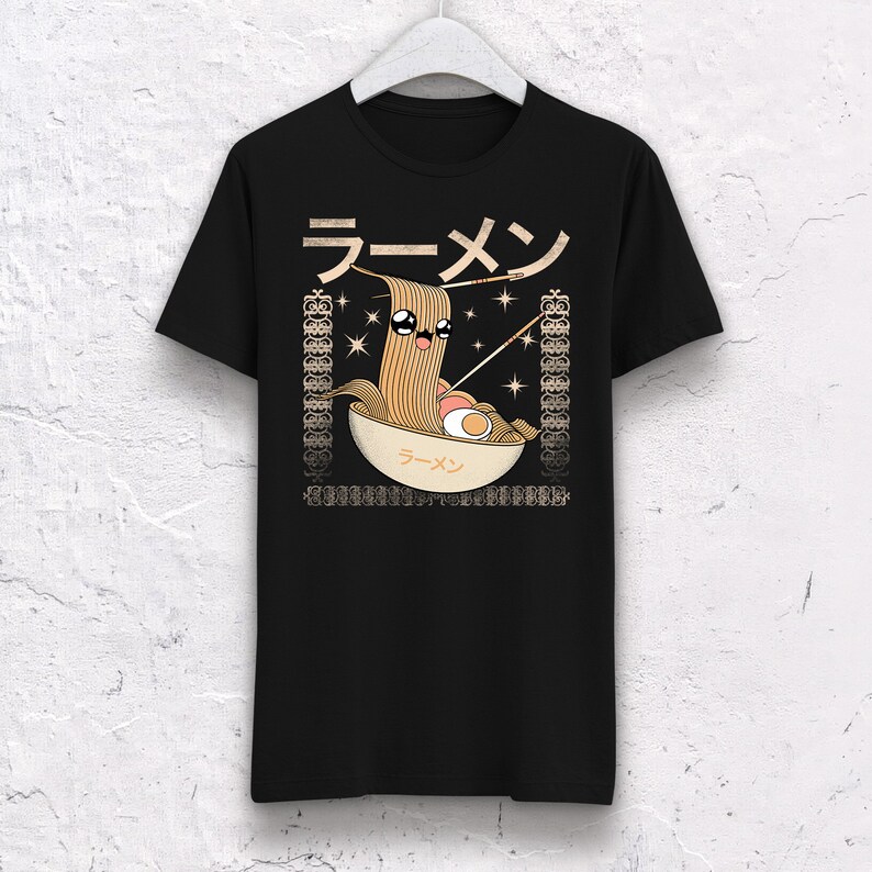 May include: Black t-shirt featuring a cartoon ramen bowl design. The illustration includes a smiling noodle character, chopsticks, and the Japanese word for ramen. The design is framed by decorative borders.
