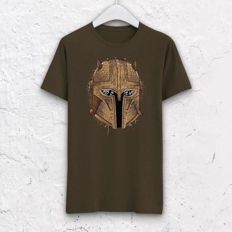 May include: A brown t-shirt with a graphic of a Mandalorian helmet. The helmet is brown and has the words "This is the way" written on it.