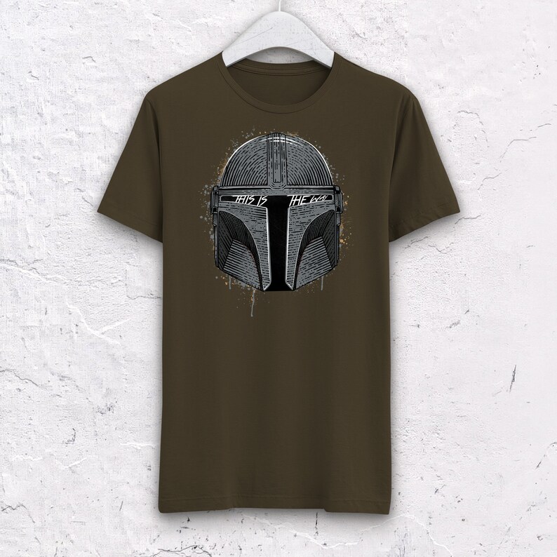 May include: A brown t-shirt with a gray and black graphic of a Mandalorian helmet. The text "This is the Way" is printed on the helmet.