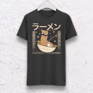 May include: Dark gray t-shirt featuring a cartoon ramen bowl with a smiling noodle character. The design includes Japanese text and decorative borders. The shirt is hanging on a white hanger.