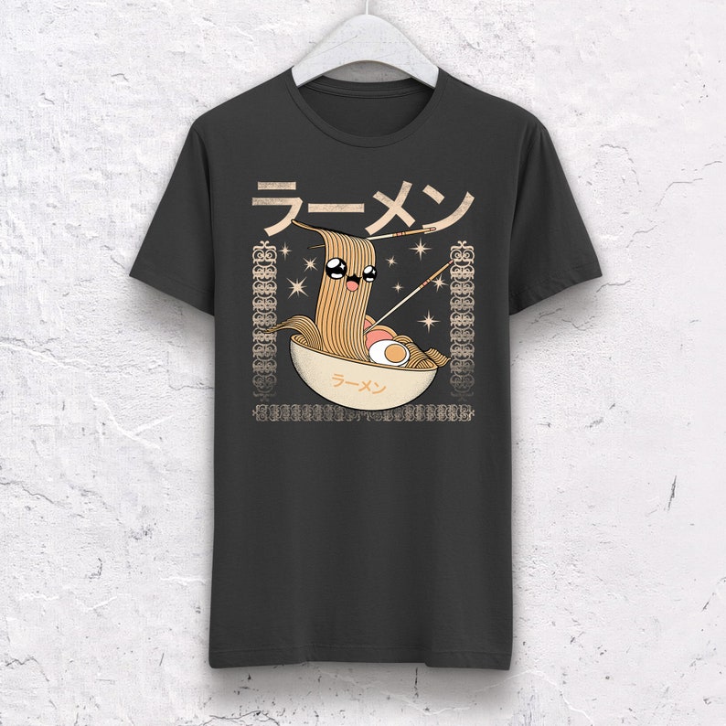 May include: Dark gray t-shirt featuring a cartoon ramen bowl with a smiling noodle character. The design includes Japanese text and decorative borders. The shirt is hanging on a white hanger.