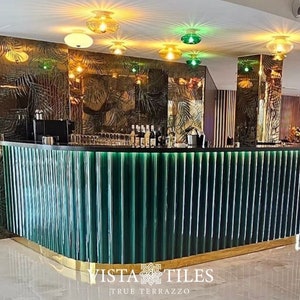 May include: A bar counter with a green tiled front and a gold trim. The counter is lit with a row of decorative lights. The background features a tropical-themed wallpaper with palm leaves.