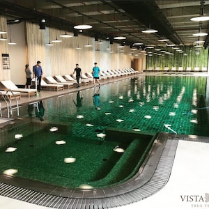 May include: A green tiled indoor swimming pool with a smaller hot tub area. The pool is surrounded by white lounge chairs and a wooden deck. The pool is located in a modern building with a high ceiling and large windows.