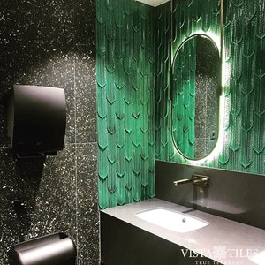 May include: A modern bathroom with a black and white speckled tiled floor. The wall behind the sink is covered in green tiles with a geometric pattern. A white porcelain sink with a gold tap is on a dark grey countertop. A large oval mirror with a gold frame is mounted on the wall. The text "Vista Tiles True Terrazzo" is visible in the bottom right corner of the image.