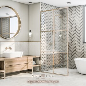 以下が含まれることがあります： ヘリンボーン柄の白い地下鉄タイル、白いバスタブ、大きな丸い鏡、そして白いシンクが付いた木製の洗面台がある、モダンなバスルーム。シャワーは、金色のアクセントが付いたガラス製の囲いがあります。画像の下部には、「VISTA TILES True Terrazzo beyond fashion for you」というテキストがあります。