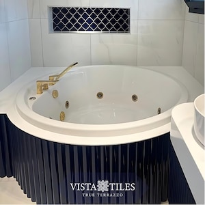 Puede incluir: Una bañera de hidromasaje blanca de forma ovalada con grifos dorados. La bañera está empotrada en una encimera blanca y una base azul marino con líneas verticales. Encima de la bañera hay un diseño decorativo de azulejos azul marino y blanco. El logotipo de Vista Tiles es visible.
