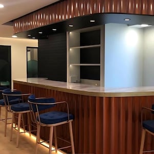 Könnte beinhalten: Ein moderner Barbereich mit geschwungener Theke und passenden Barhockern. Die Bar hat eine braune, vertikal geriffelte Fassade mit goldenen Zierleisten. Die Hocker haben blaue Samtsitze und goldene Rahmen. Einbaustrahler beleuchten den Raum.