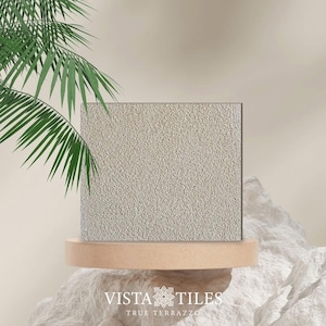 Op de afbeelding: Een close-up van een lichtgrijze terrazzo tegelstaal. De tegel heeft een gespikkelde uitstraling met kleine witte en grijze chips. De tegel wordt gepresenteerd op een houten platform.