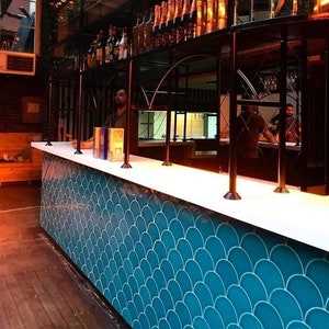 May include: A bar counter with a white countertop and a teal blue tiled backsplash. The tiles are arranged in a fish scale pattern. The bar is lit from below, creating a warm glow.
