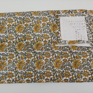 May include: A white fabric with a yellow and green floral pattern. The fabric has a small pocket sewn into the top right corner.