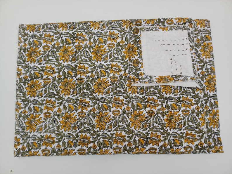 May include: A white fabric with a yellow and green floral pattern. The fabric has a small pocket sewn into the top right corner.