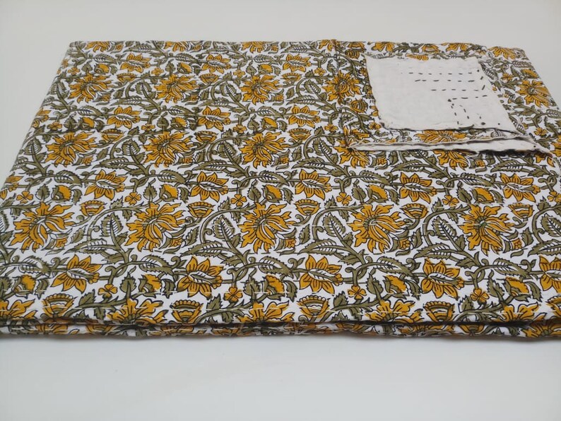 May include: A white cotton fabric with a floral pattern in shades of yellow and green. The fabric is folded in thirds and a small white square of fabric is visible in the upper right corner.