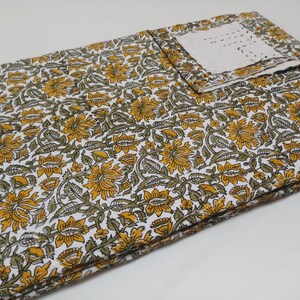 May include: A white cotton fabric with a yellow and green floral print. The fabric is folded in half and has a small white square of fabric on top.