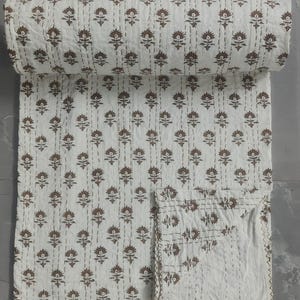 May include: A rolled-up white quilt with a repeating brown floral pattern. The quilt is stitched with white thread and has a brown border. The floral design is evenly spaced across the white background. The quilt is folded over, showing the pattern.