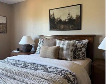 May include: A bedroom scene featuring a wooden bed frame with a headboard, adorned with pillows in various textures and patterns. The bed is covered with a white duvet and a gray patterned blanket. A framed landscape painting hangs above the bed, and a lamp sits on a nightstand.
