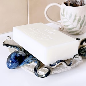 May include: A ceramic soap dish with a blue and white octopus design. The dish holds a white rectangular bar of soap. The octopus has a glossy finish, and the dish has a leaf-like shape.