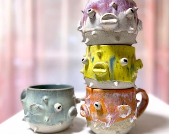May include: Four ceramic mugs shaped like pufferfish, each with a different color glaze. The mugs are stacked on top of each other, with the largest mug on the bottom and the smallest mug on top. The mugs are all decorated with white eyes and a mouth.