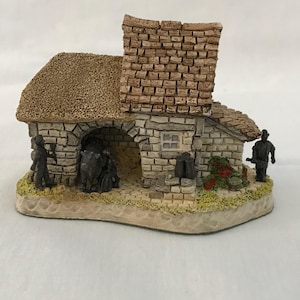 May include: A miniature stone cottage with a thatched roof and brick chimney. The cottage features an arched entrance, a small window, and miniature figures of people. The base is painted with a grassy texture.
