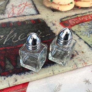 May include: Two clear glass salt and pepper shakers with silver lids sit on a decorative tray with holiday-themed designs. The tray has text that says "Joy", "cheer", and "Snowflake". The background has a red and white pattern.