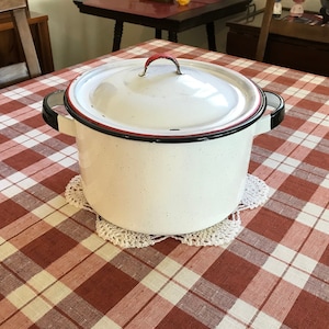 May include: White enamelware pot with a red and black rim and a lid. The pot is sitting on a white crocheted doily on a red and white checkered tablecloth.