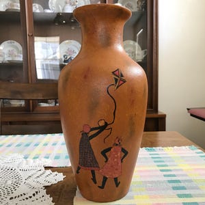 May include: A tall, brown ceramic vase with a wide base and narrow neck. The vase features a painted design of two figures flying a kite. The kite is red, black, and white. The figures are wearing dresses and coats. The vase is approximately 18 inches tall.