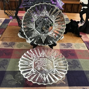 May include: Two clear, pressed-glass plates with a starburst design and a central fruit motif. The plates are on a plaid table runner with a Halloween-themed decoration in the background.