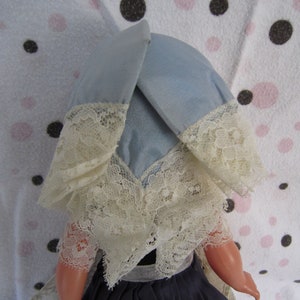 Pu&ograve; includere: Una bambola vintage con un berretto di pizzo blu e bianco. La bambola ha un viso di porcellana e indossa un vestito blu.