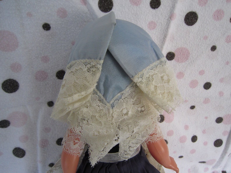 Pu&ograve; includere: Una bambola vintage con un berretto di pizzo blu e bianco. La bambola ha un viso di porcellana e indossa un vestito blu.