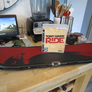 May include: A black and red skateboard controller with the game "Tony Hawk: Ride" for PlayStation 3. The controller has a red graphic design and is on a wooden surface.