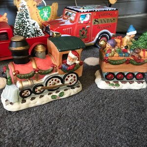 May include: A festive Christmas train set with a red engine and a car filled with gifts and a Christmas tree. The set includes a Santa Claus figure and is decorated with holly and snow.